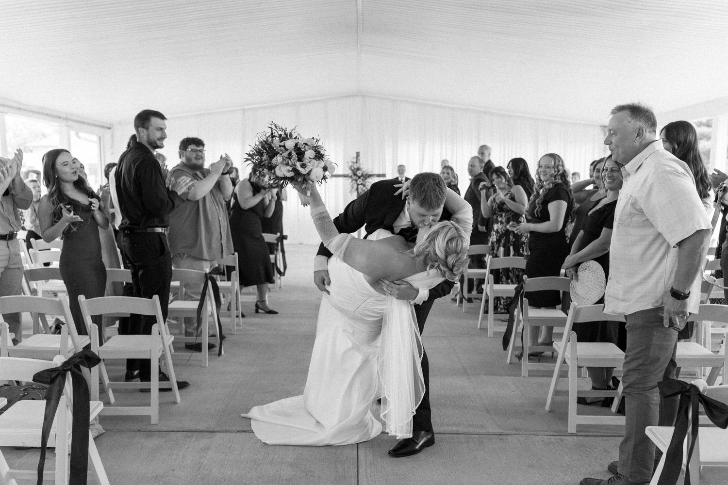 A black and white with wedding guests seated in rows of chairs under a large outdoor tent and couple dip kiss. Located in Bellbrook, Ohio.