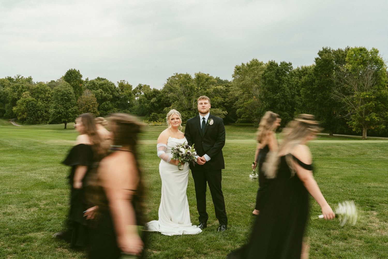 Wedding party members run and dance across a lush green field during outdoor celebration festivities. Located in Bellbrook, Ohio.