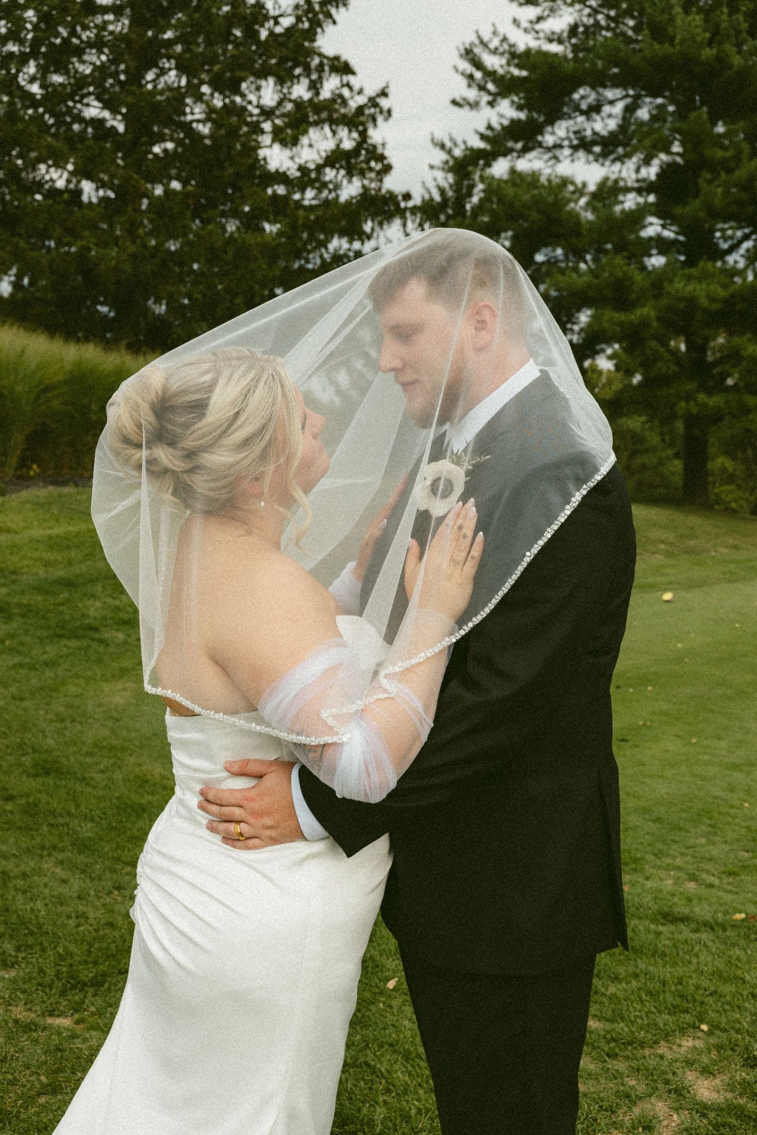 A romantic wedding moment captured outdoors with a bride in white gown and groom in black suit sharing an intimate moment under a veil.