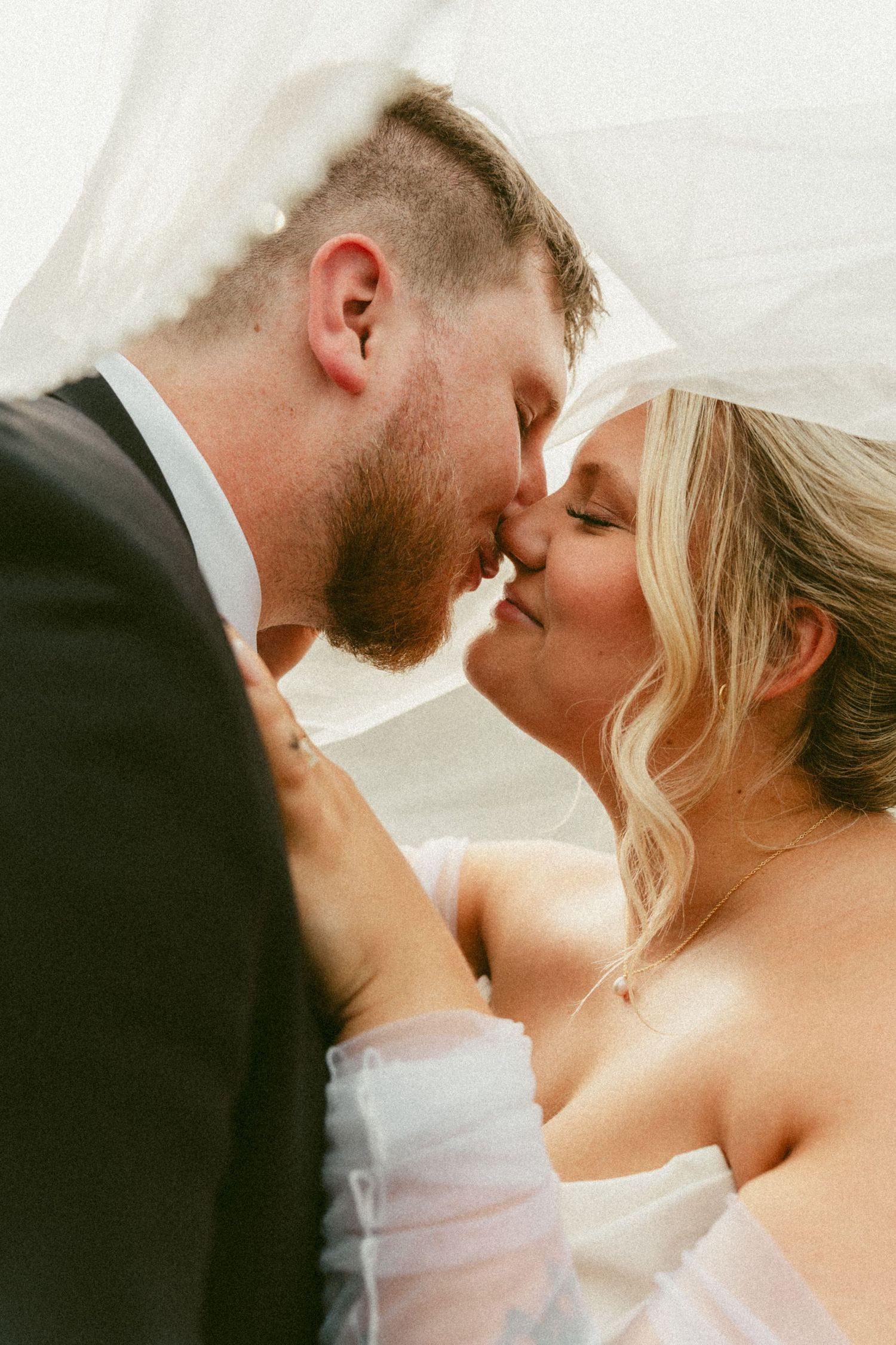 A romantic wedding couple shares an intimate kiss under flowing white fabric in soft natural lighting. Located in Bellbrook, Ohio.