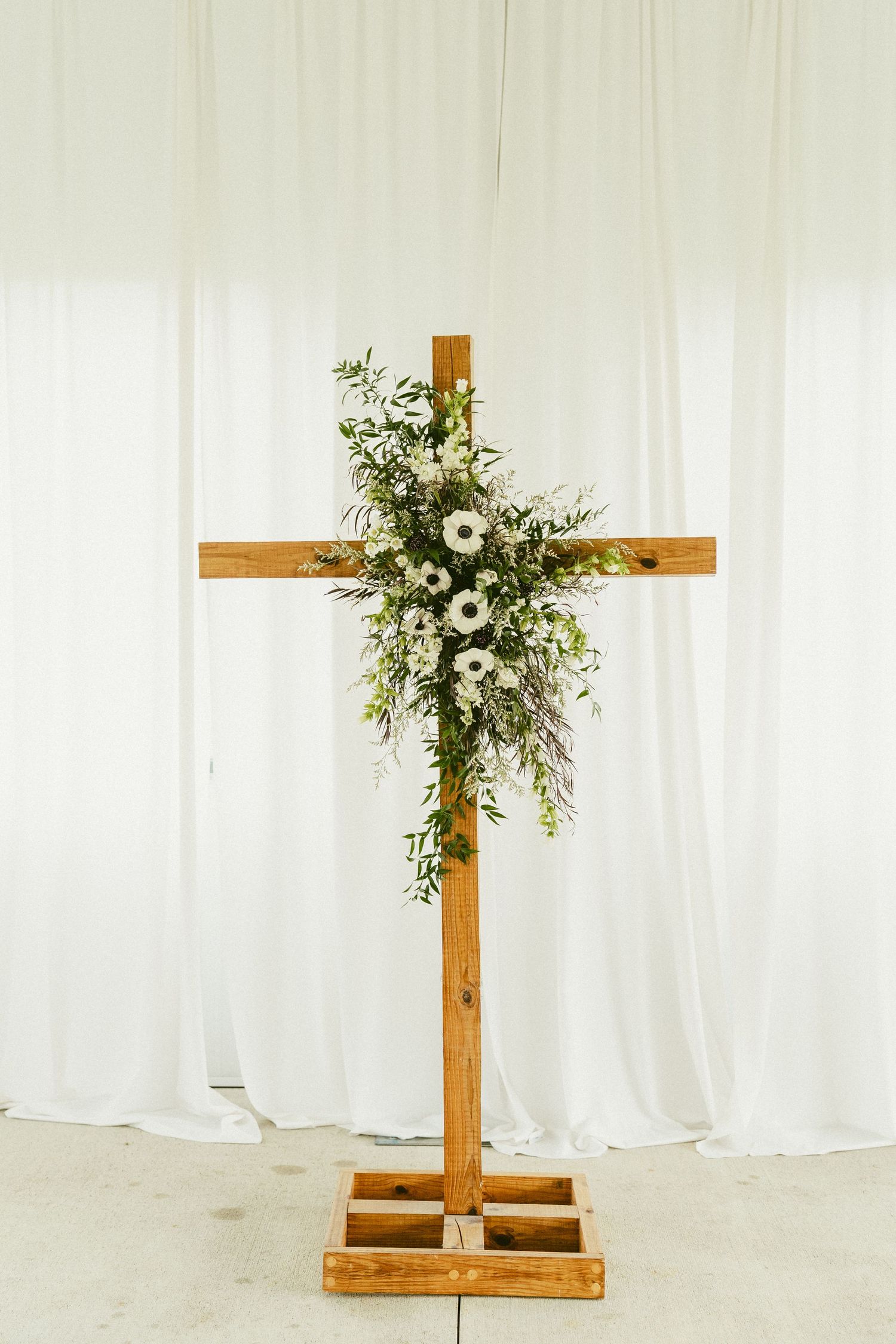 A wooden cross adorned with white floral arrangements and greenery against a light fabric backdrop. Located in Bellbrook, Ohio.