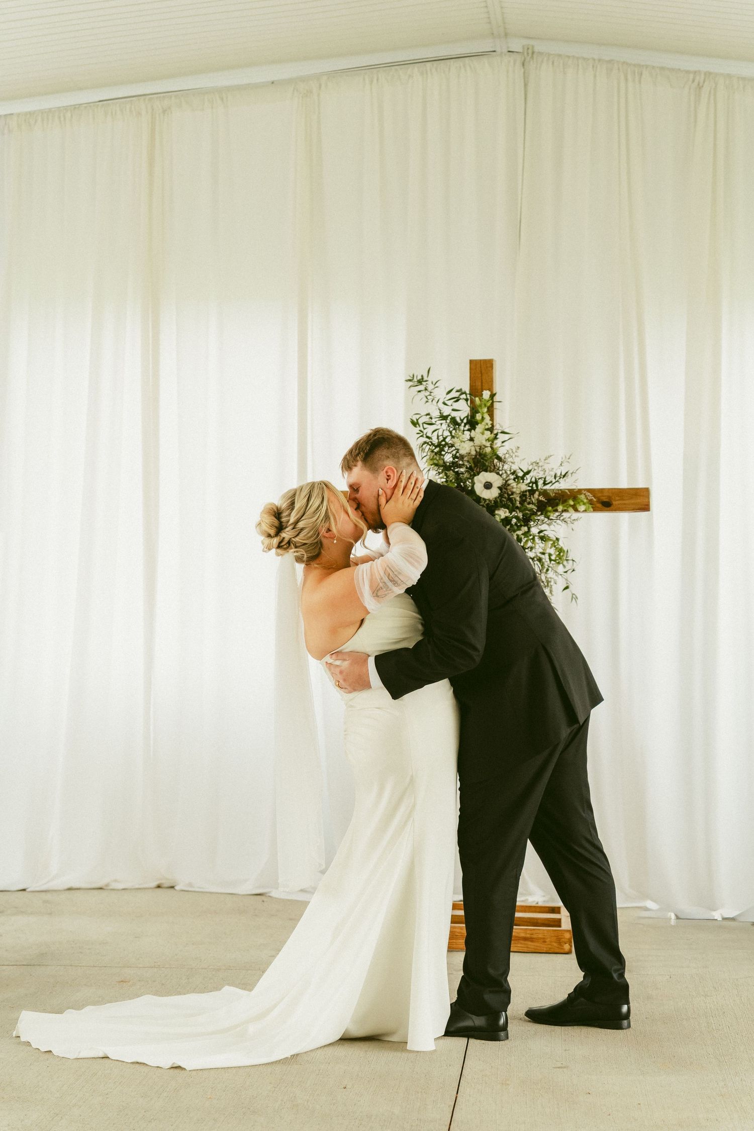 A romantic wedding couple shares a kiss against a white curtain backdrop during their ceremony. Located in Bellbrook, Ohio.