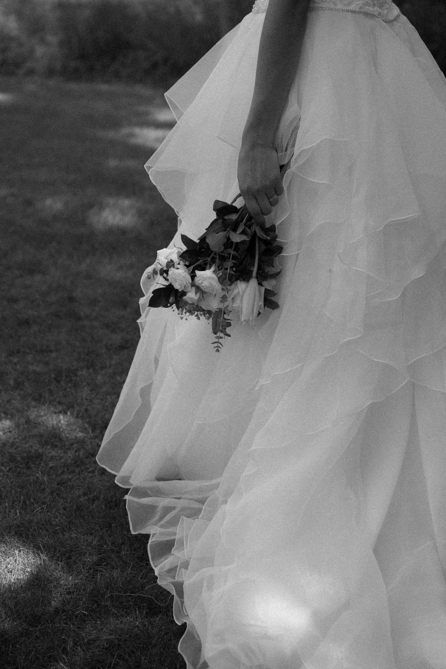 Black and white photo showing flowing layers of a white wedding dress and bouquet against a grassy background. Located in Dayton, Ohio.