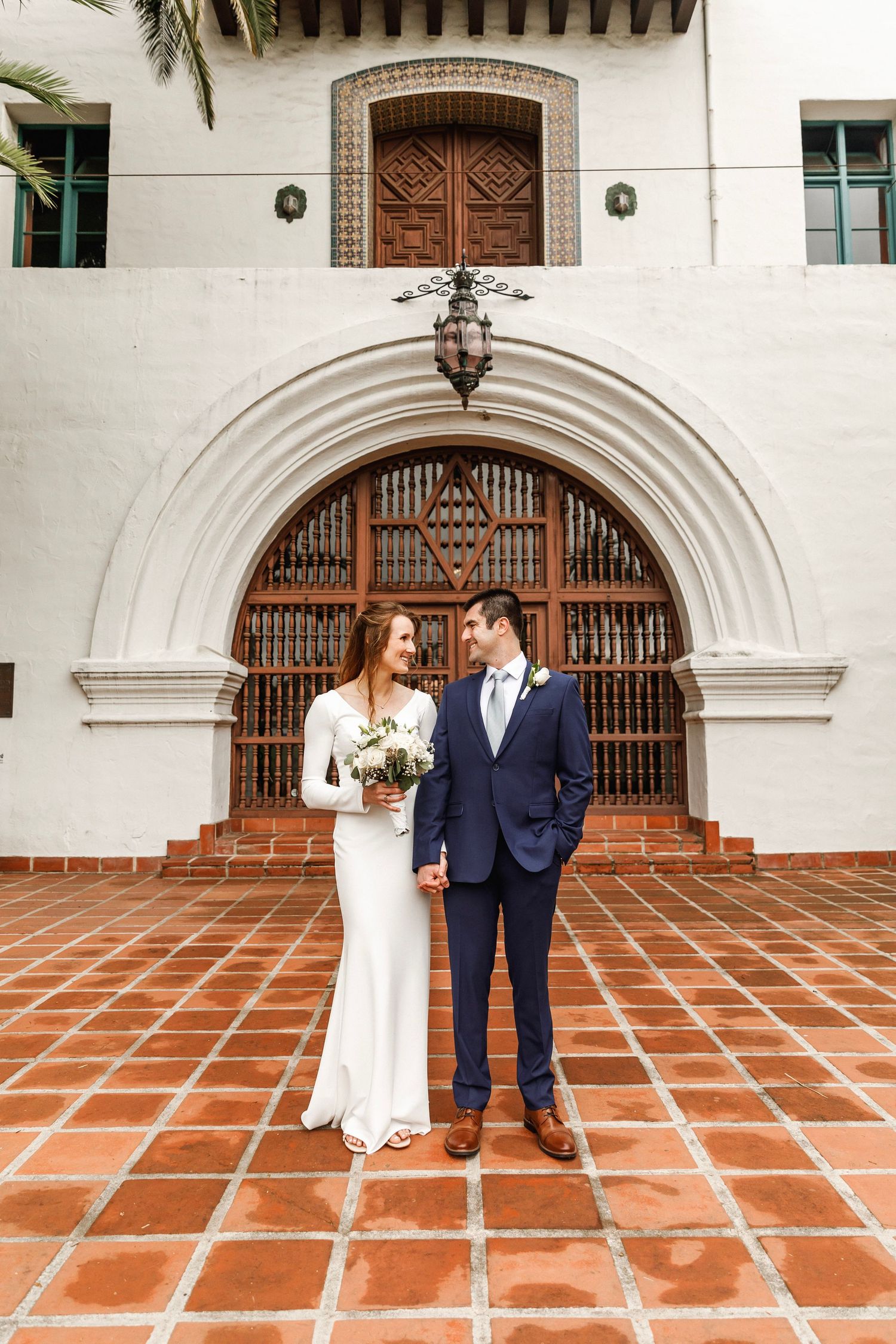 Bride and groom smiling at each other before ornate arched doorway of a Spanish-style building.