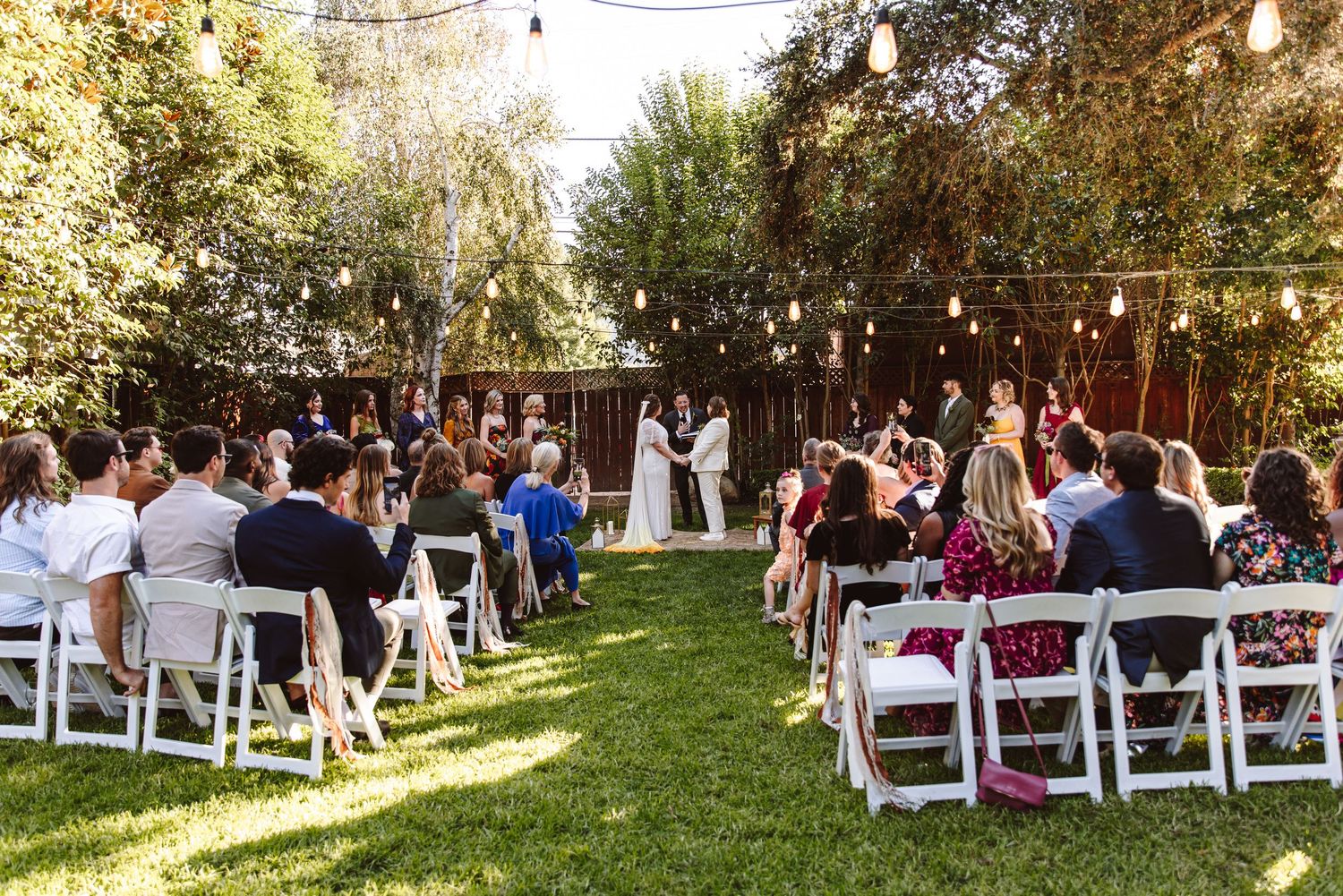 Outdoor backyard wedding ceremony with guests seated on white chairs under string lights.