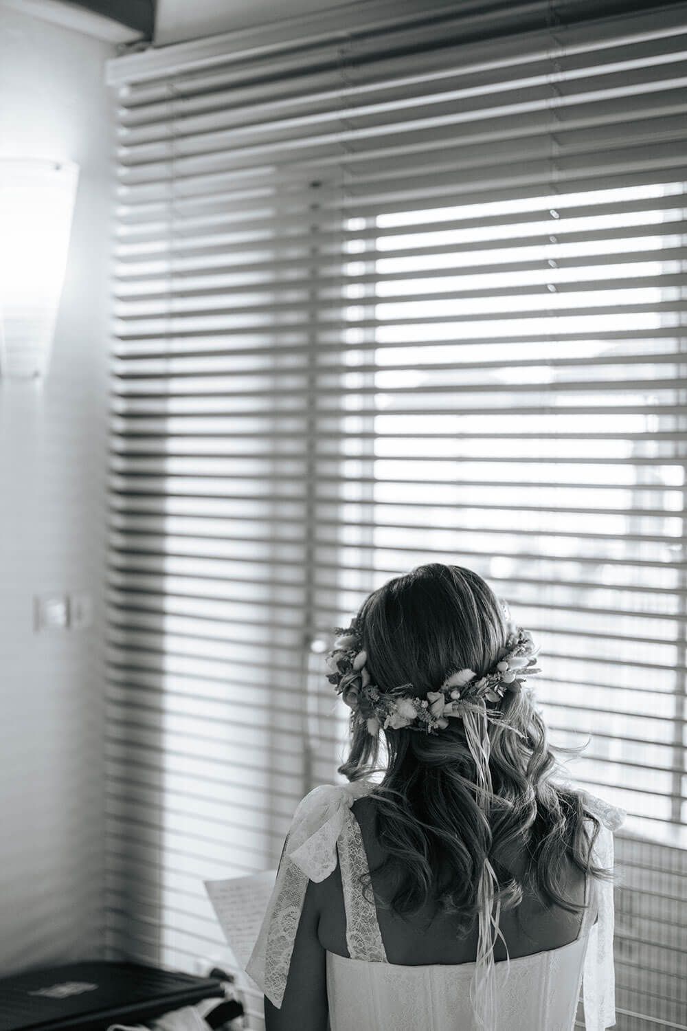A black and white photo showing a person with flowers in their hair sitting by a window with venetian blinds.
