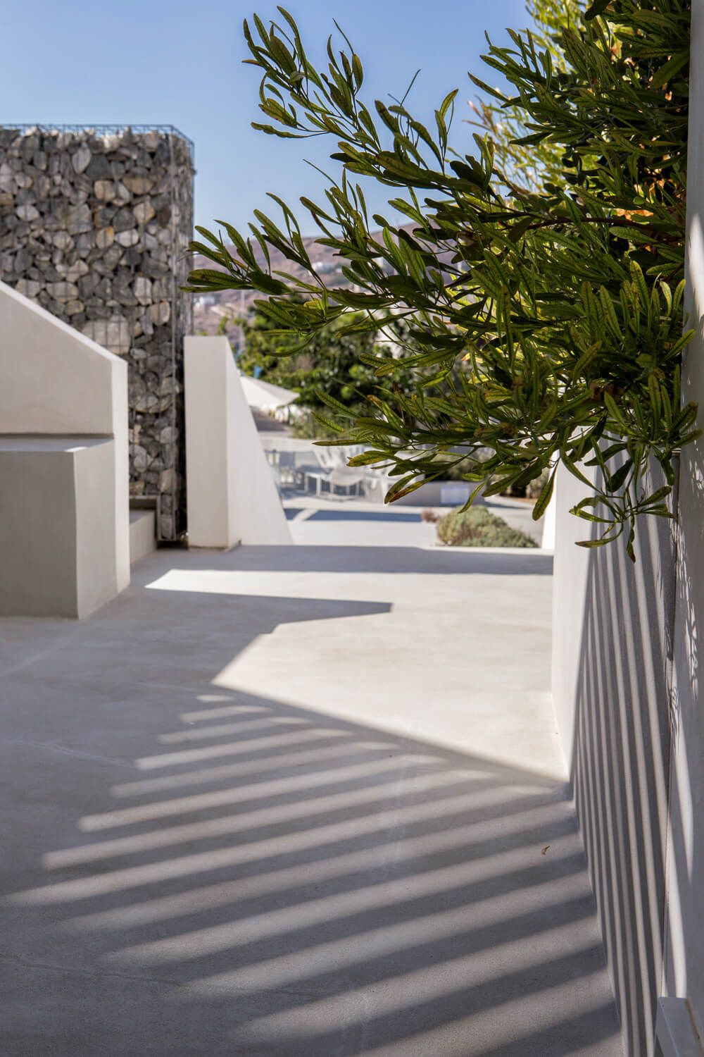Modern Mediterranean terrace with stone wall, white planters and olive branches casting dramatic shadows on concrete floor.