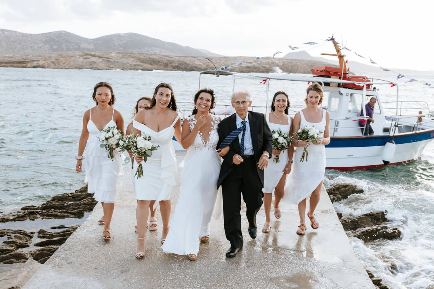 Bride with bridesmaids and father in white dresses walking along a seaside pier with a boat behind.