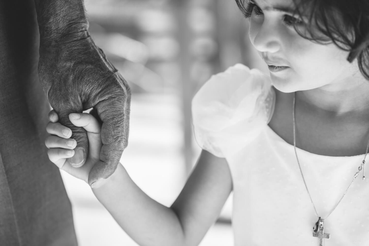 Black and white photo of a child holding an elderly person's hand, wearing a cross necklace.