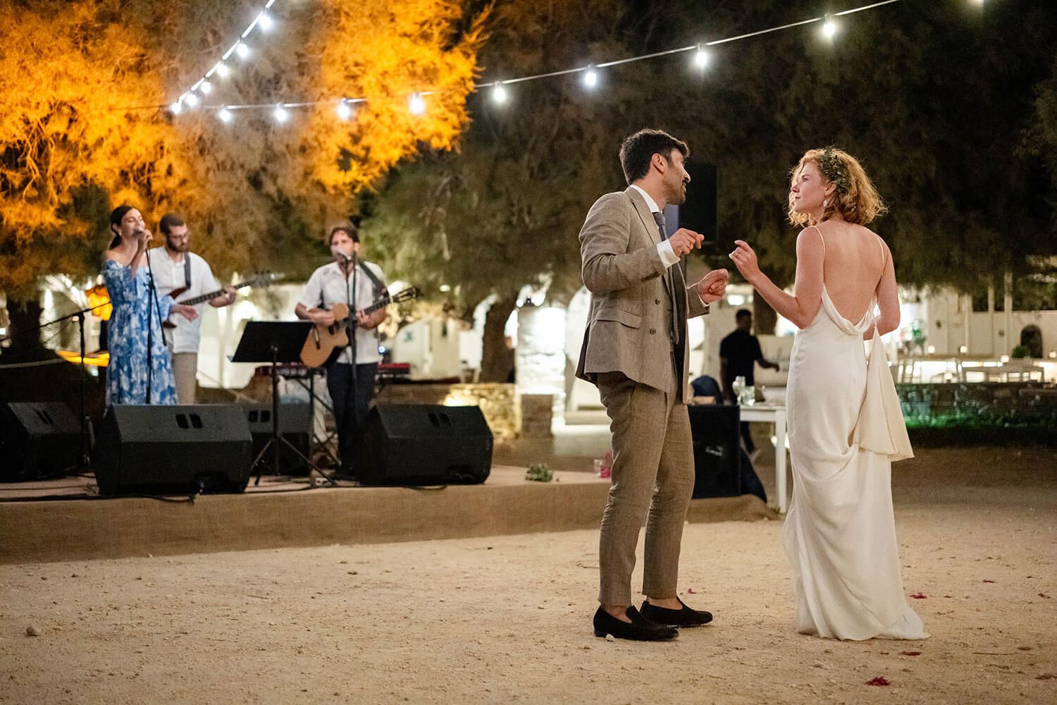 A romantic outdoor wedding reception at night with string lights illuminating couples dancing on a sandy dance floor in Paros Greece