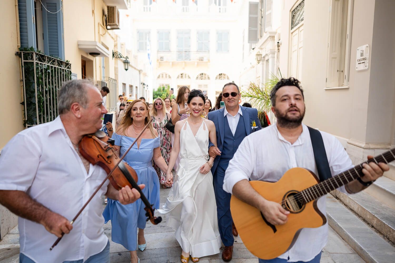 Bride walk through the streets of Syros with local musicians during outdoor wedding celebration.