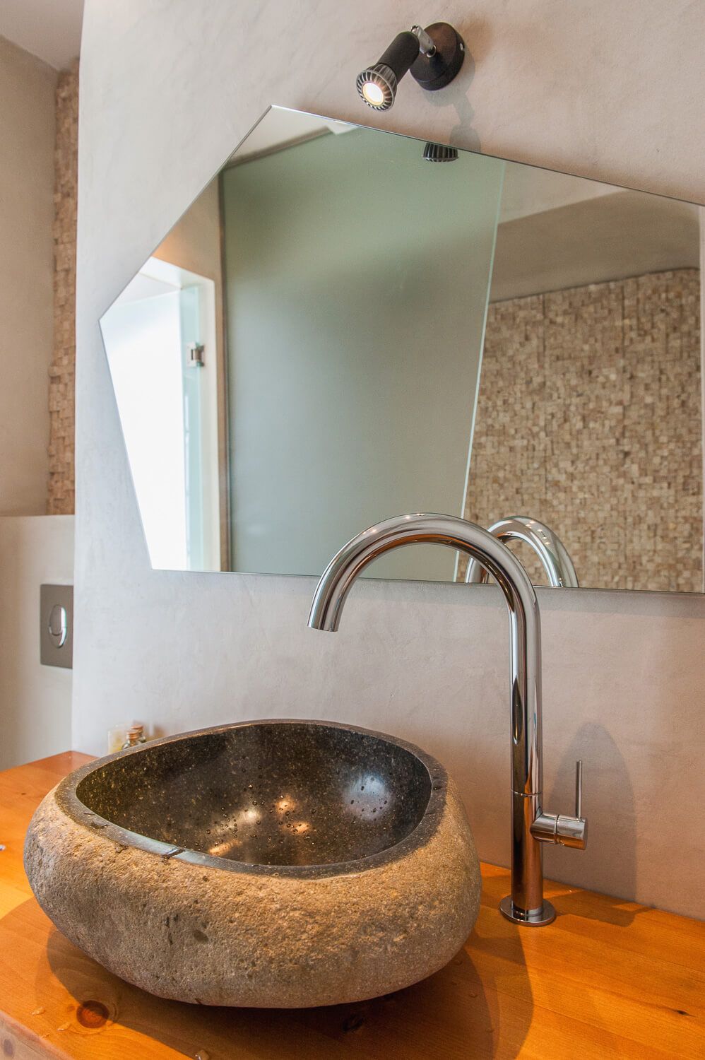 Modern bathroom vanity with stone vessel sink, wooden countertop and angular wall mirror with spotlight fixture.