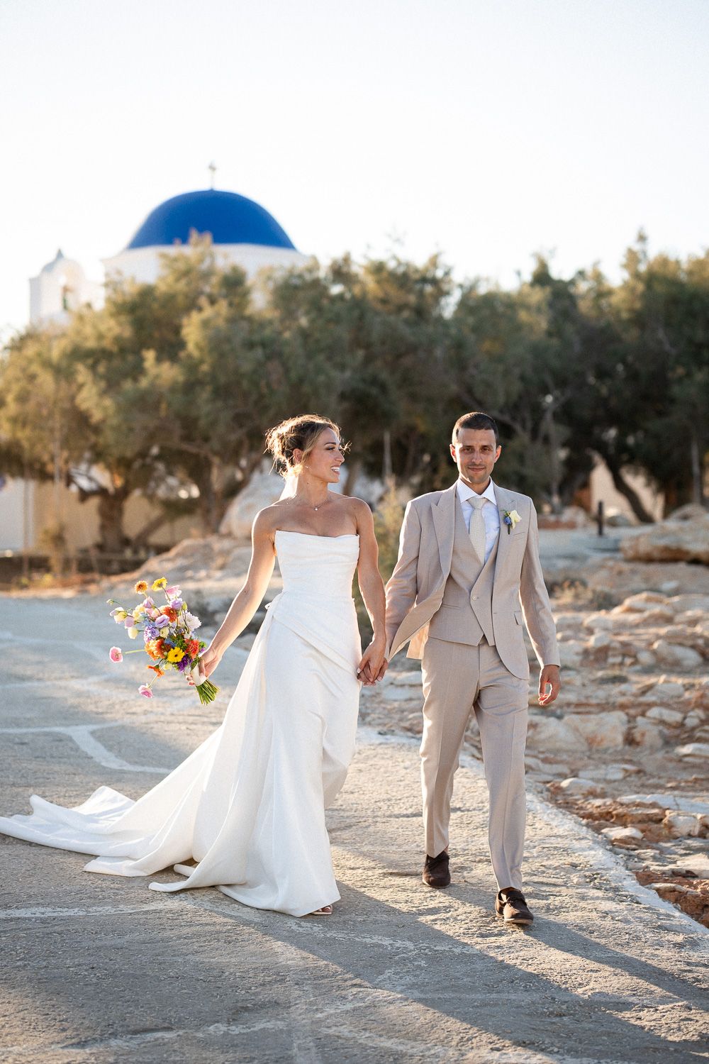 Newlyweds walk with Agios Fokas in the distance, showcasing authentic Paros wedding photography.