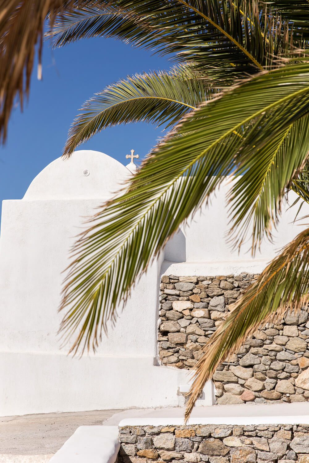Palm fronds frame a whitewashed church wall with stone foundation against bright blue sky in Mediterranean style.