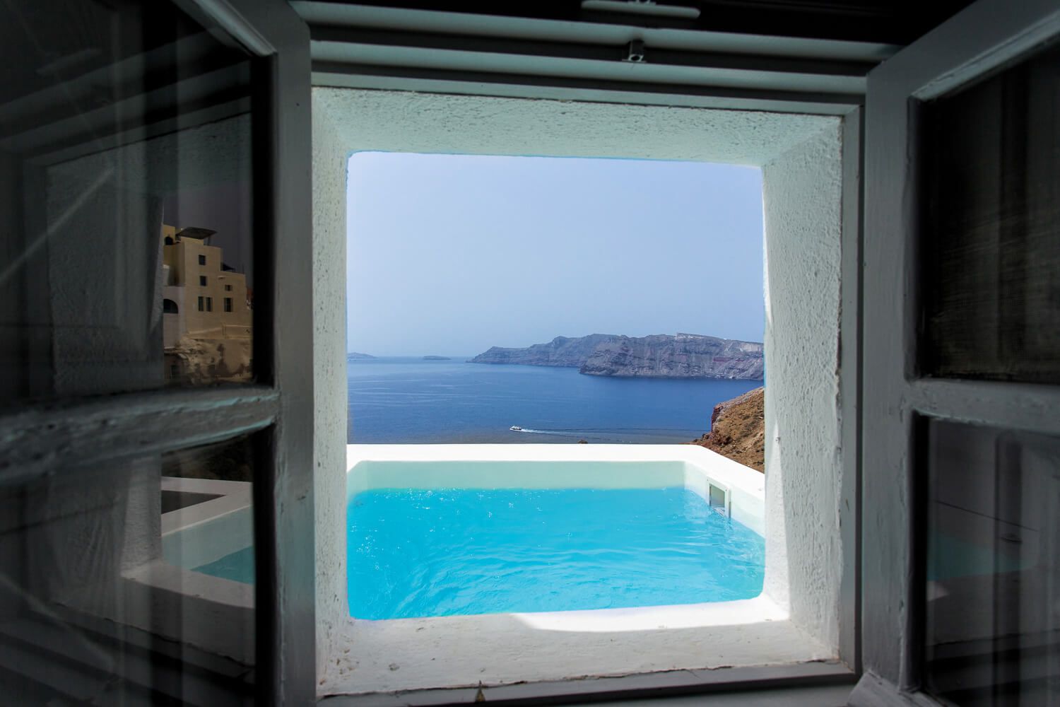 View through open window of infinity pool overlooking Santorini caldera and Aegean Sea cliffs.