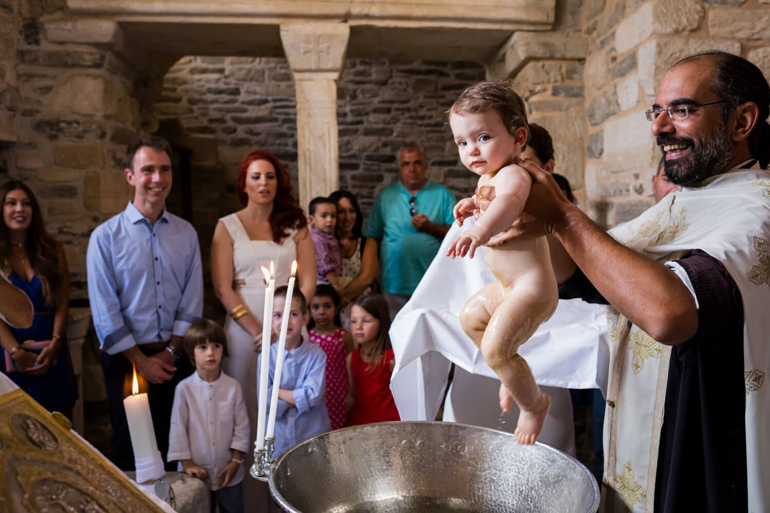 Priest holds baby over baptismal font during christening ceremony with family watching in Paros.