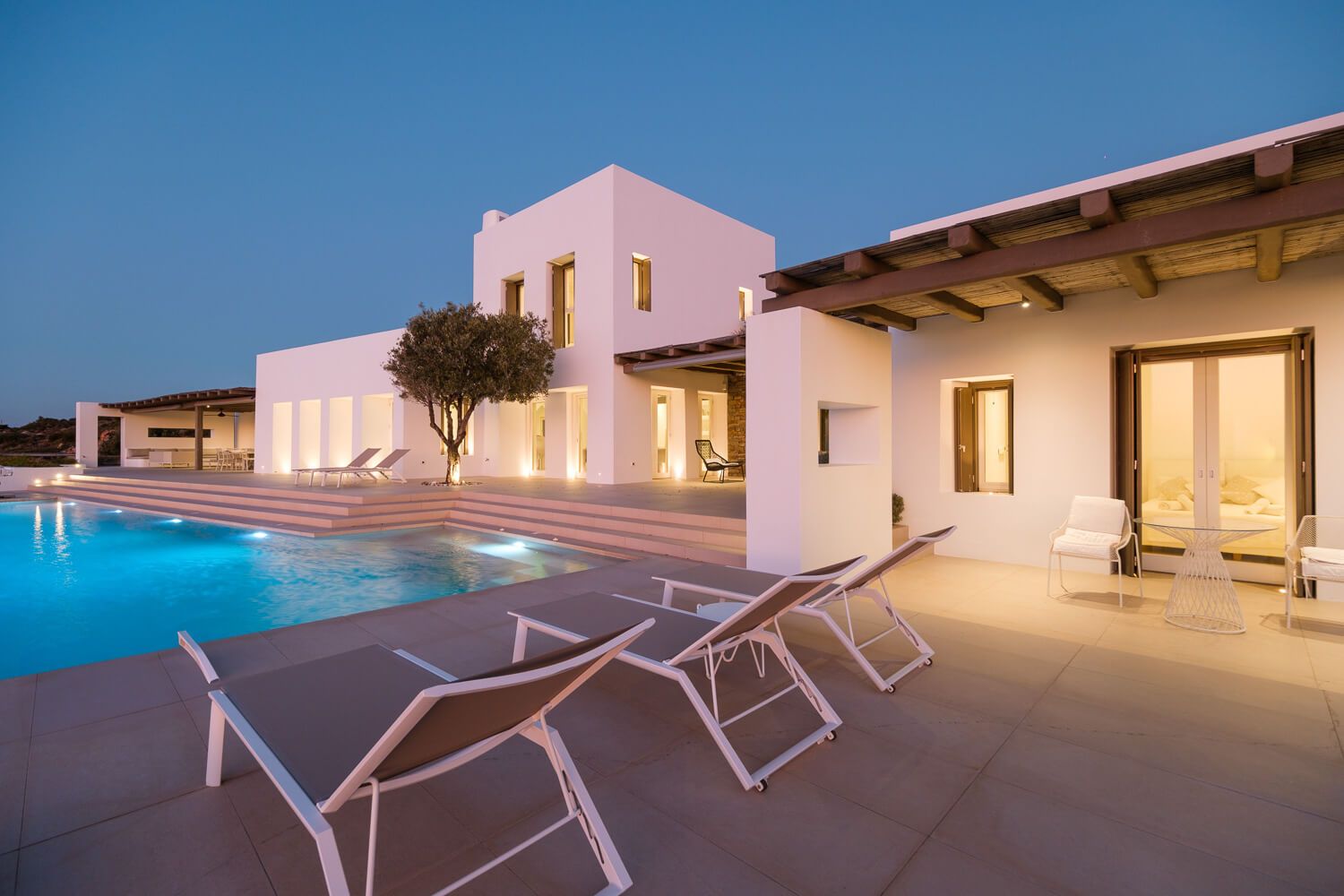 Luxurious modern white villa with illuminated swimming pool and lounge chairs at dusk in Mediterranean style in Paros