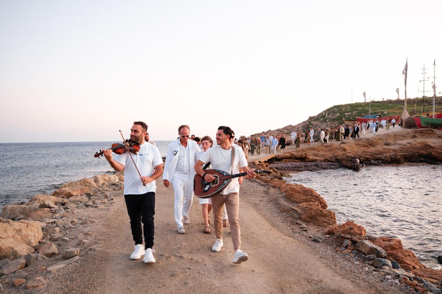 Musicians playing violin and guitar lead wedding procession along scenic coastal path at sunset.