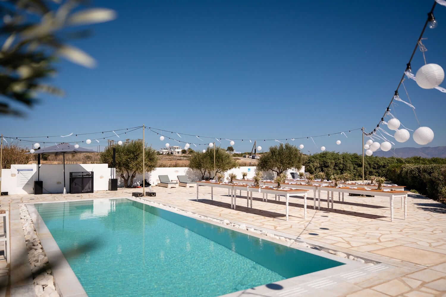 A turquoise swimming pool surrounded by white lounge chairs and string lights at a Mediterranean-style resort.