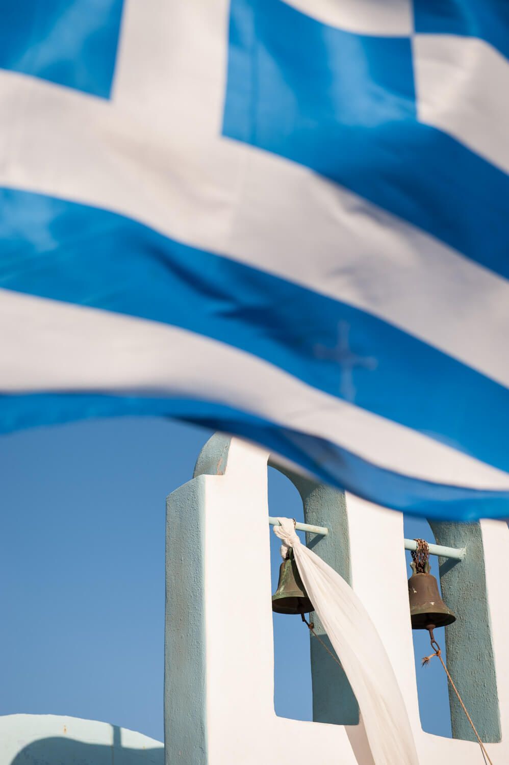 Greek flag waving over a white church bell tower with two bronze bells against a clear blue sky.