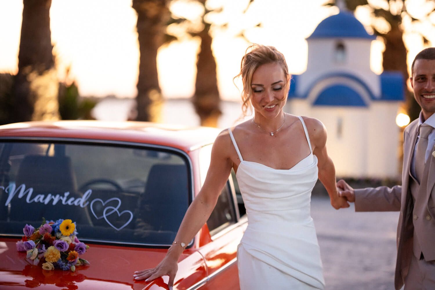 Smiling bride in white dress beside red car with Married sign at Palmtree Events Paros.