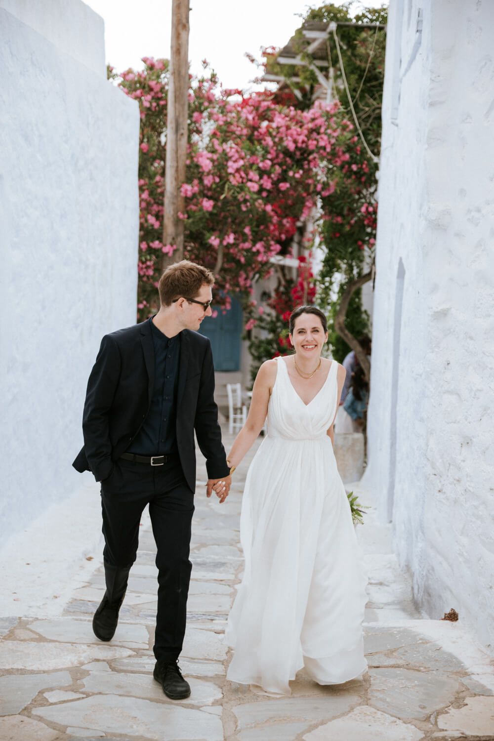 Bride and groom walk hand in hand through charming whitewashed Amorgos alley with pink bougainvillea