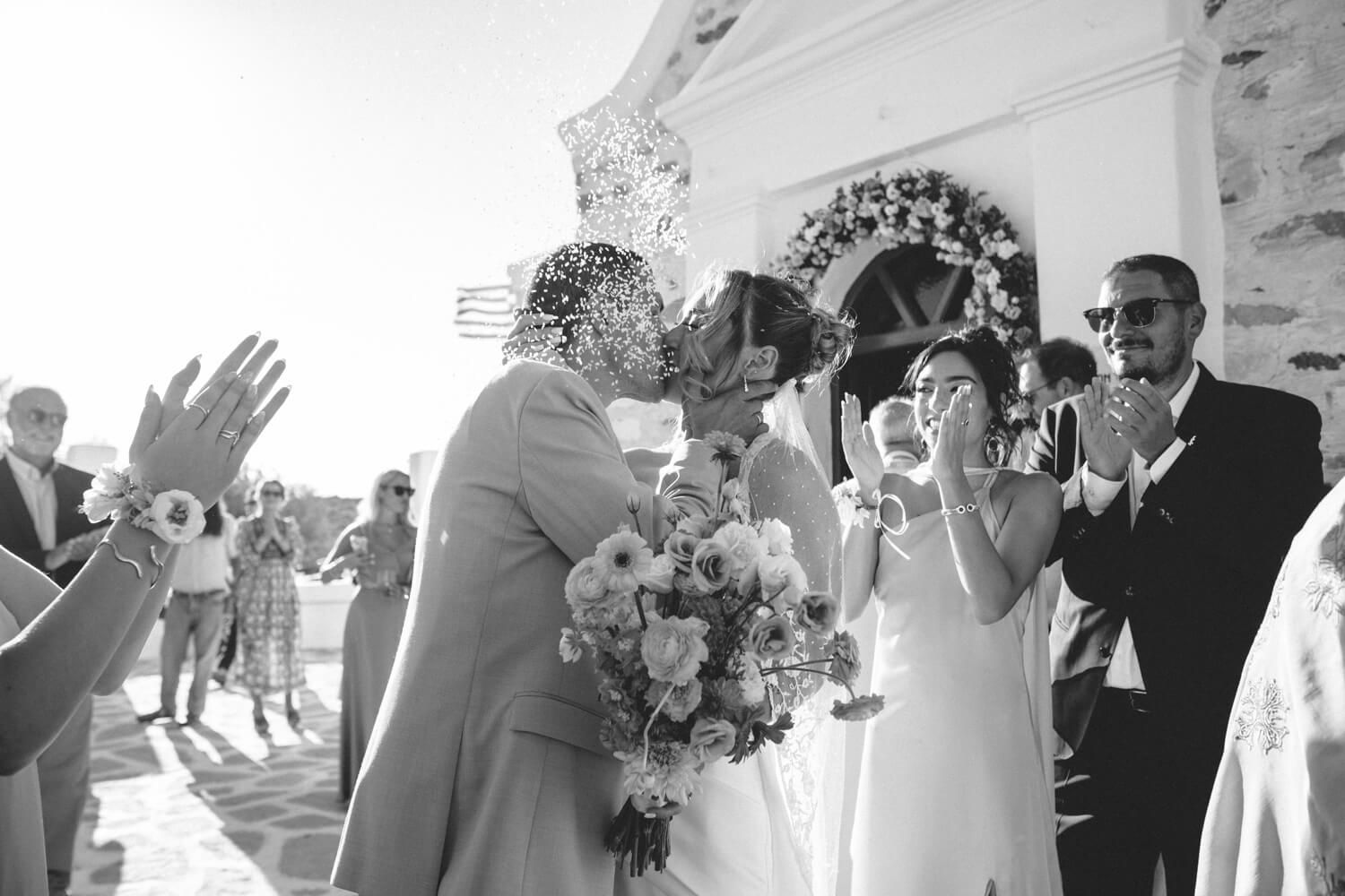 Newlyweds share a kiss outside a Greek chapel as guests applaud in a black and white wedding photo.