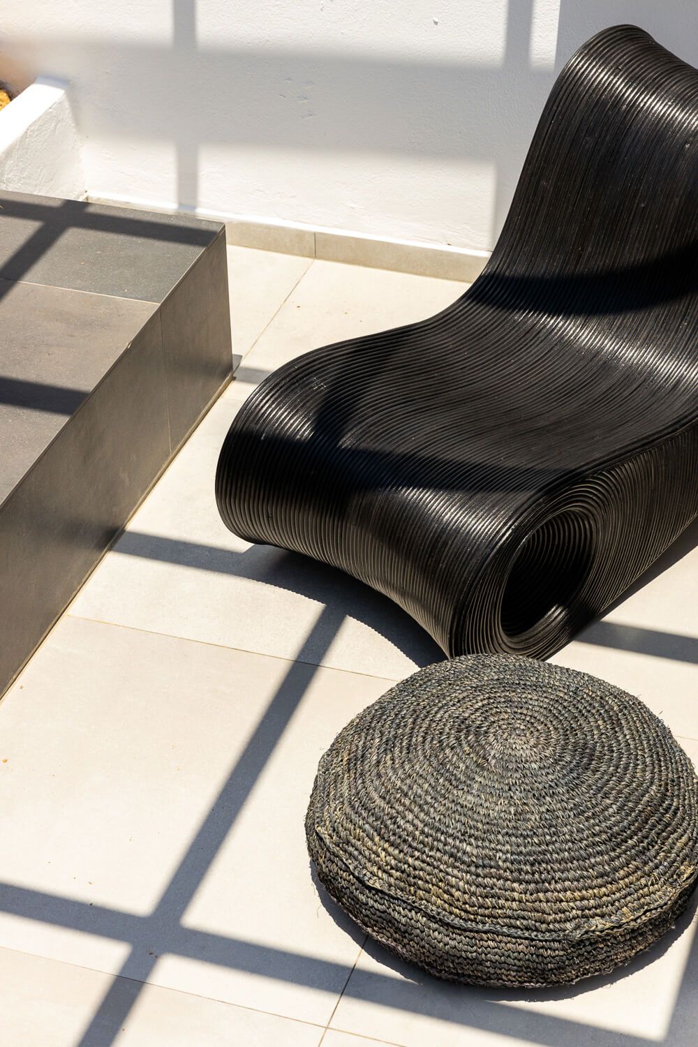 Modern black wavy rattan chair beside a round woven pouf on white tiles in a sunlit outdoor patio.