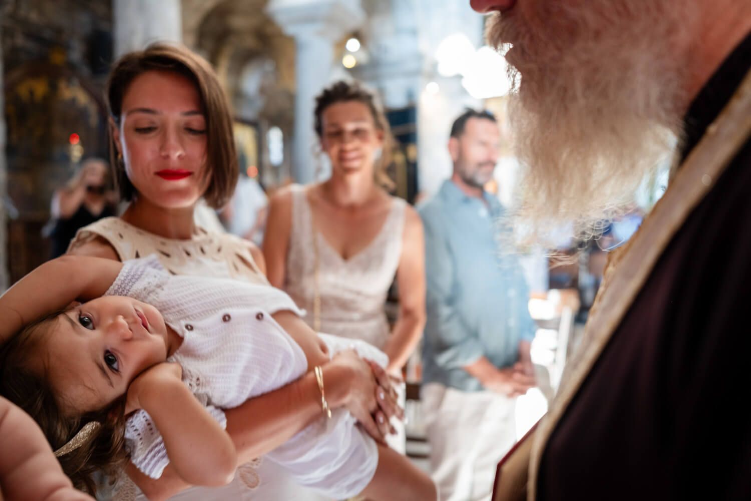Woman holding baby in white dress during a religious baptism ceremony with priest and guests.