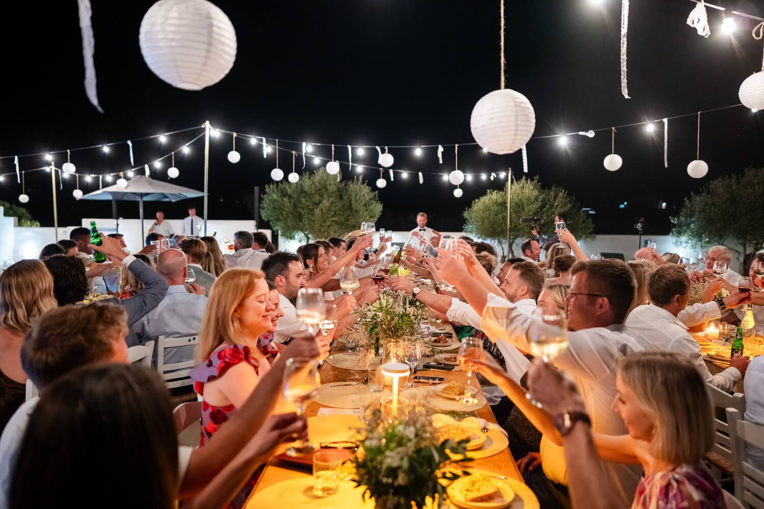 A long outdoor dining table illuminated by hanging paper lanterns and string lights creates a festive evening atmosphere.