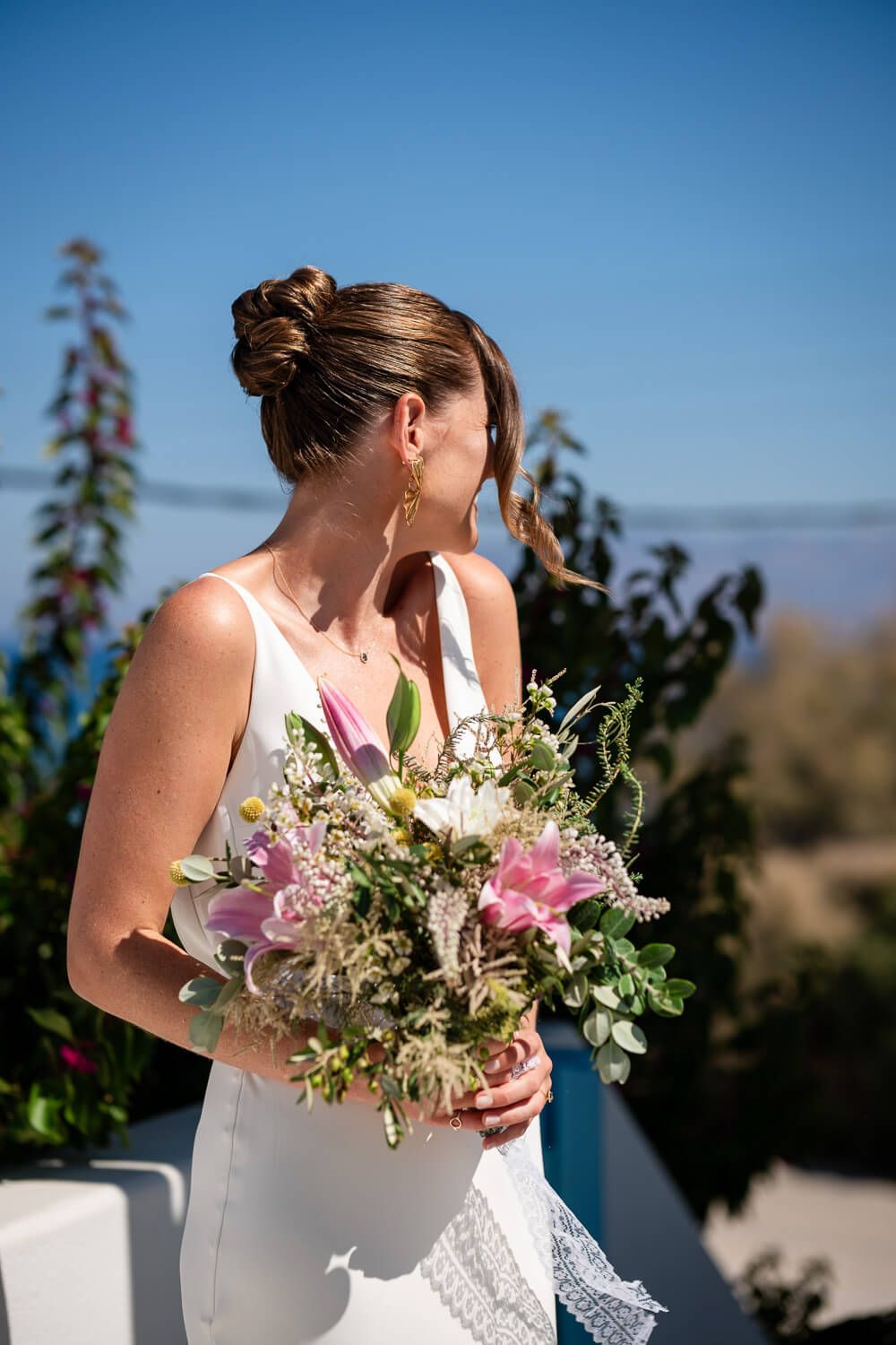 Bride in white dress holds a beautiful pink and white floral bouquet against a bright blue sky backdrop in Paros Greece
