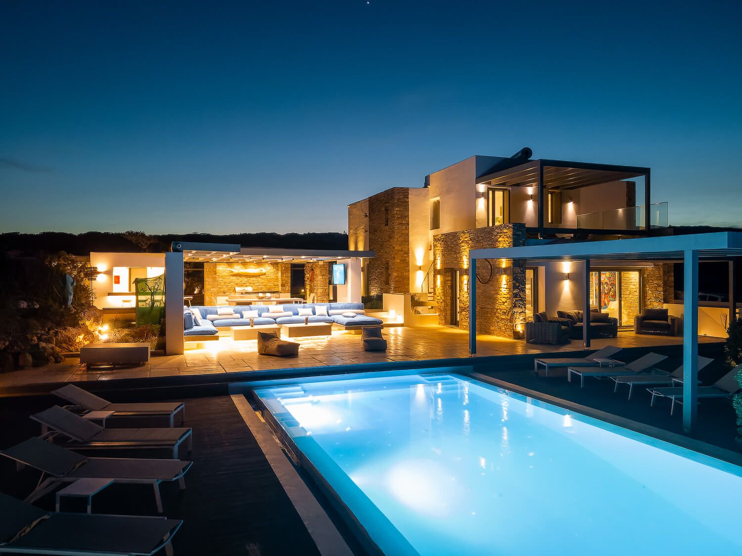 Illuminated luxury resort swimming pool and villa glowing at dusk with warm ambient lighting and calm blue water.