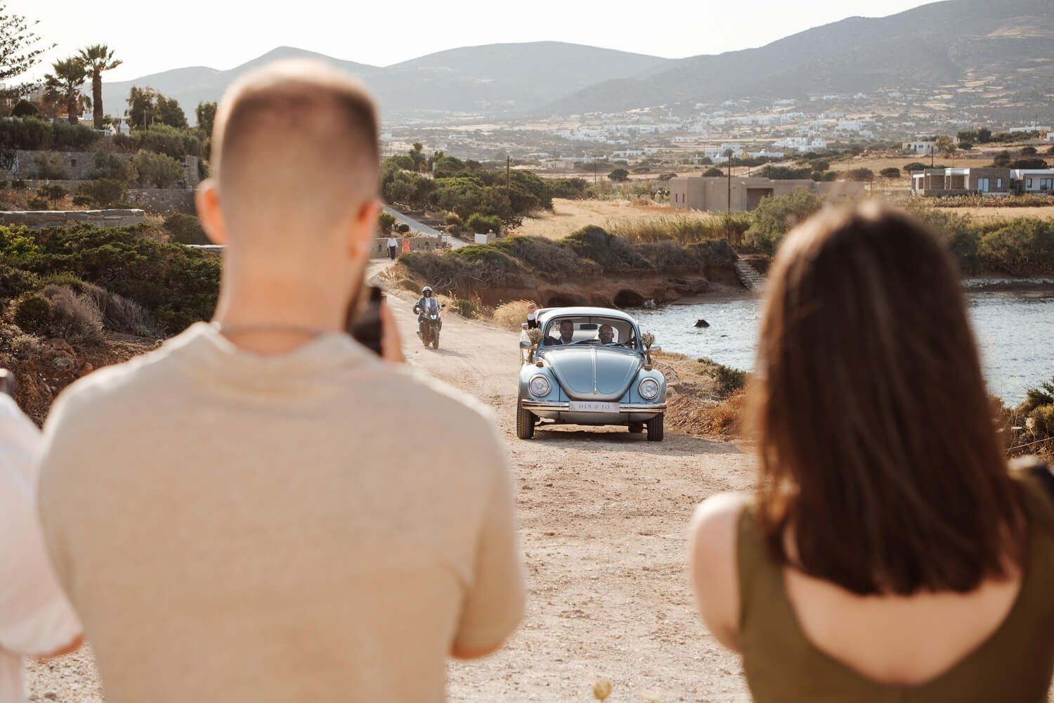 Groom with his best man arriving at Pyrgaki chapel in a vintage VW Beetle while guests watching