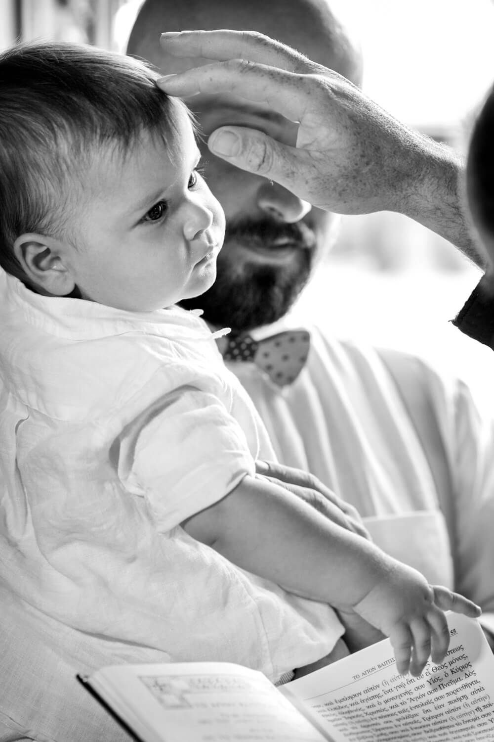 B&W photo of a baby in white outfit being blessed by a priest during a baptism ceremony in Andros.