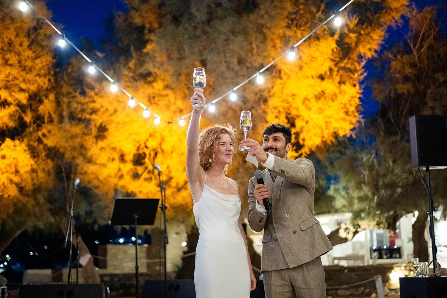 Wedding couple dances under twinkling lights with autumn trees glowing orange in the background at night.