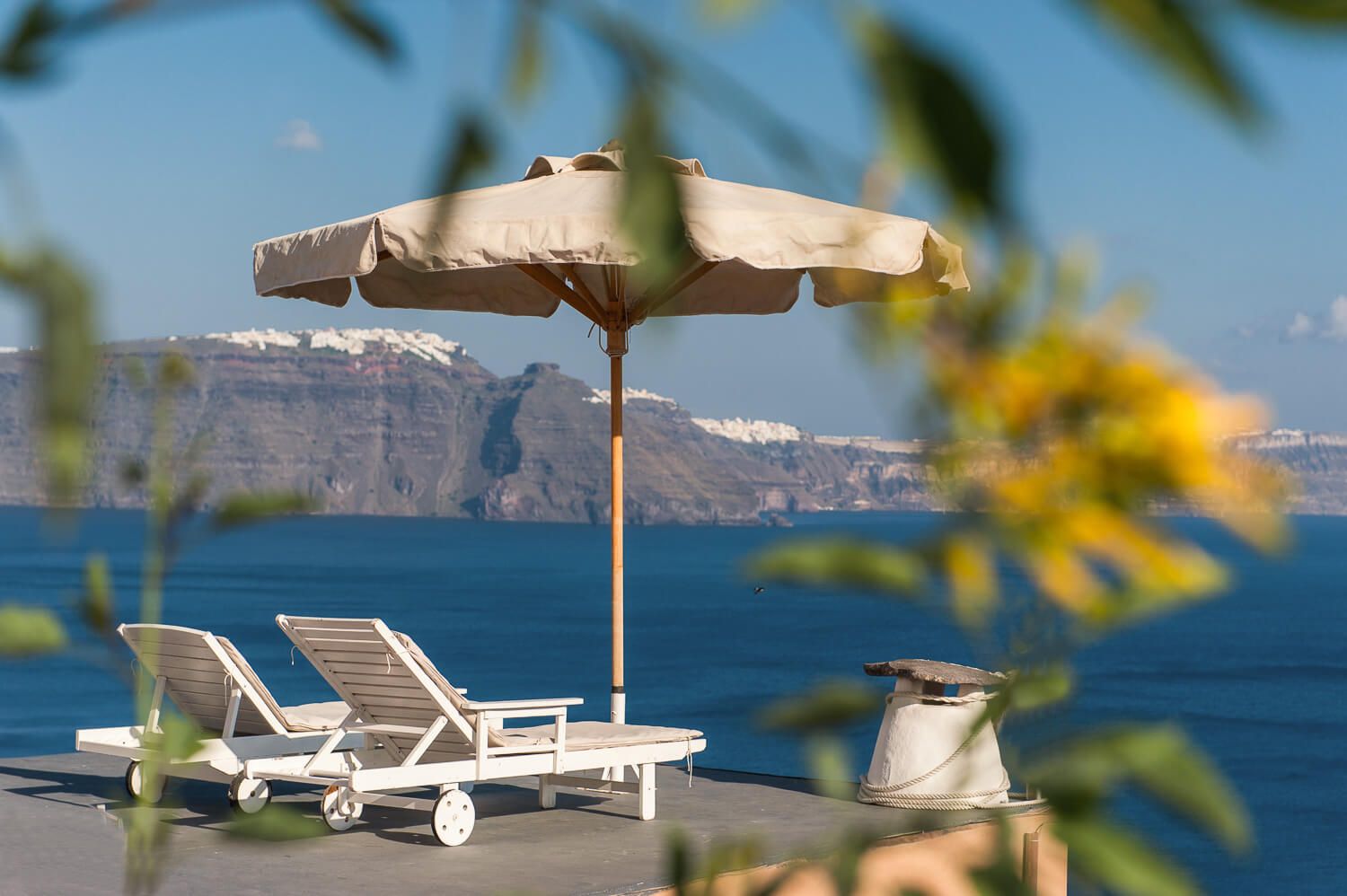 White lounge chairs and umbrella on a terrace overlooking the blue Mediterranean Sea with cliffside views in Santorini.