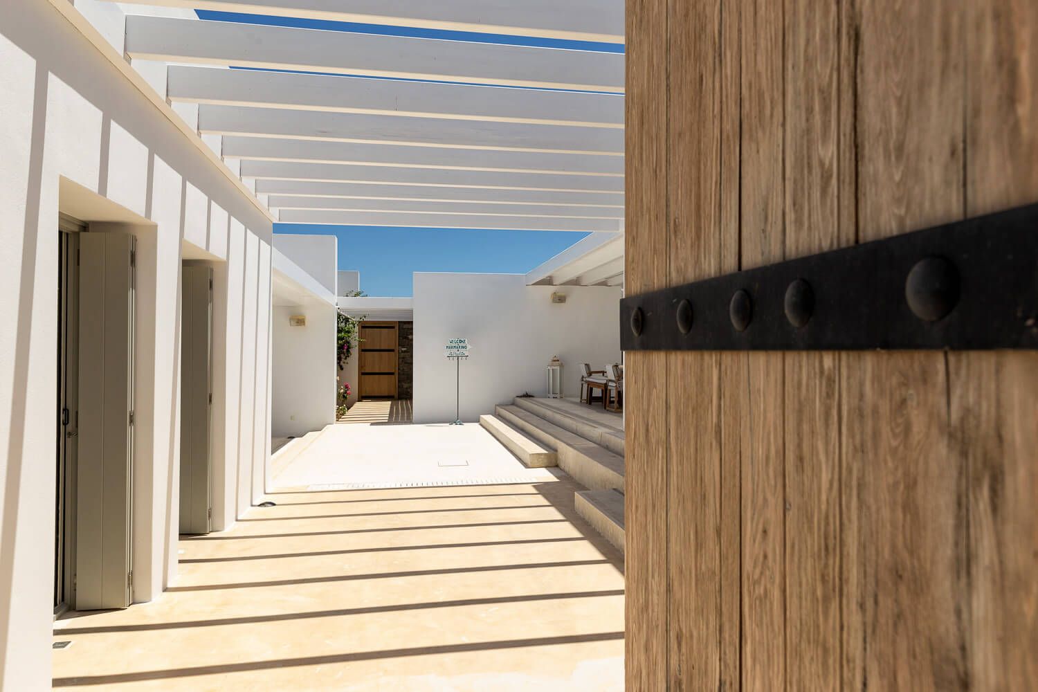 Modern white courtyard with wooden pergola, sandy pathway, and minimalist architecture in Paros.
