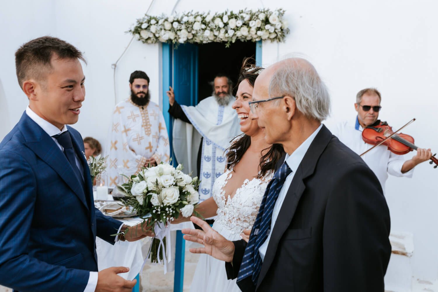 Groom in blue suit greets elderly man as bride in white lace dress holds bouquet outside Greek church wedding.