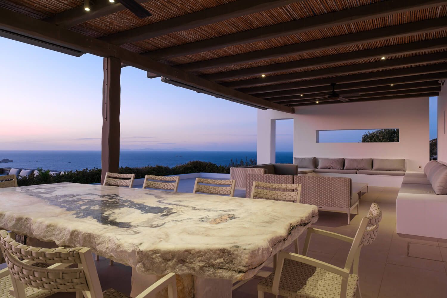 A luxurious beachfront terrace with white columns and wooden beams overlooks a coastline at sunset.