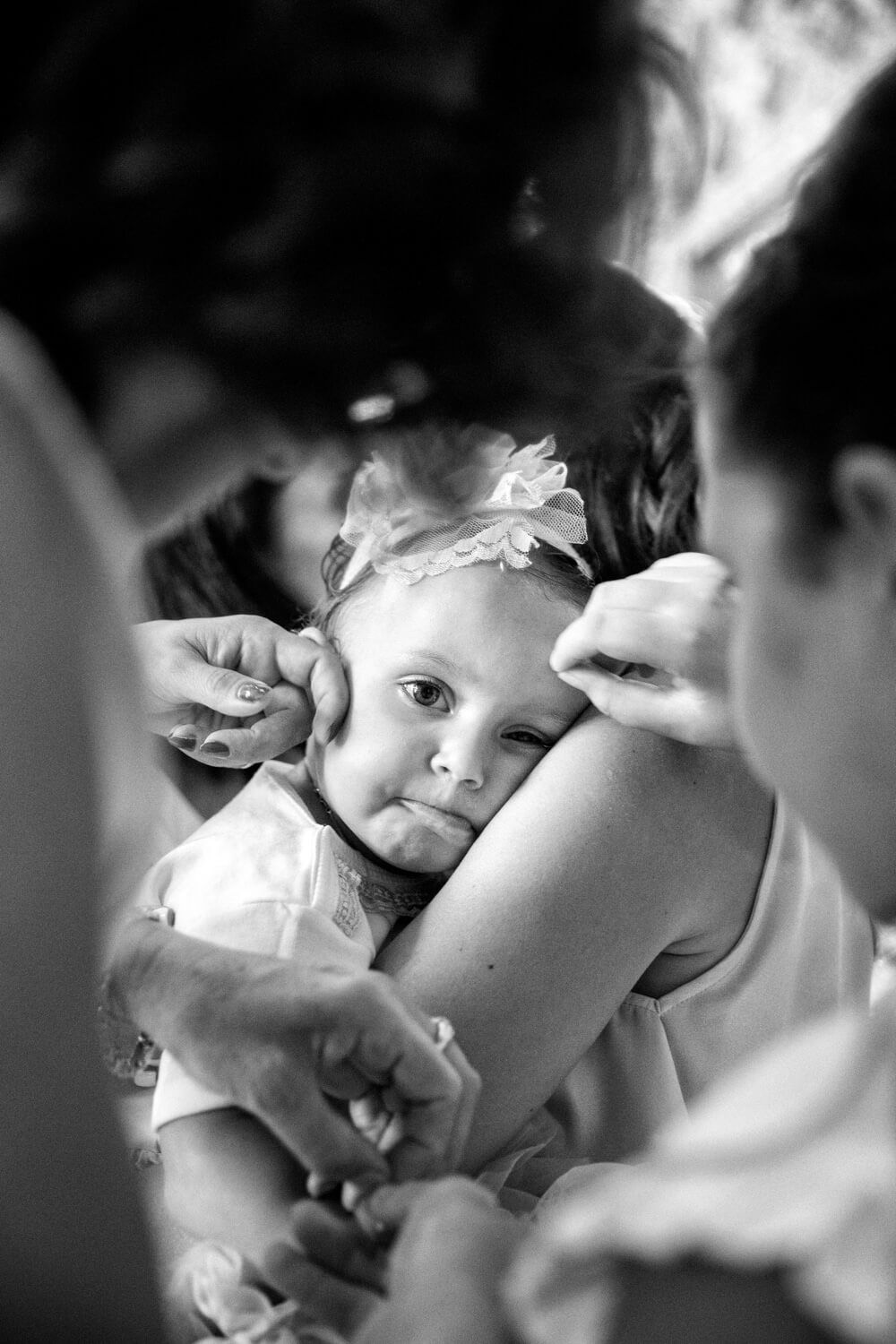 Black and white photo of a baby girl with flower headband being hugged by adults at a Greek baptism.
