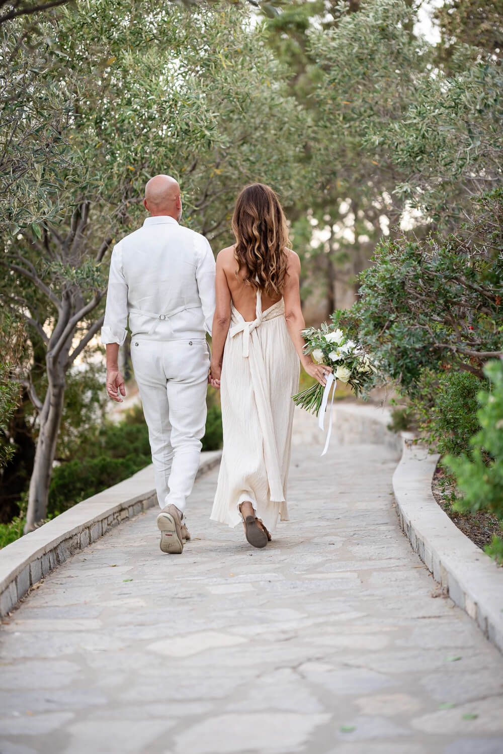 A couple in white attire walks hand in hand down a stone pathway surrounded by lush greenery.