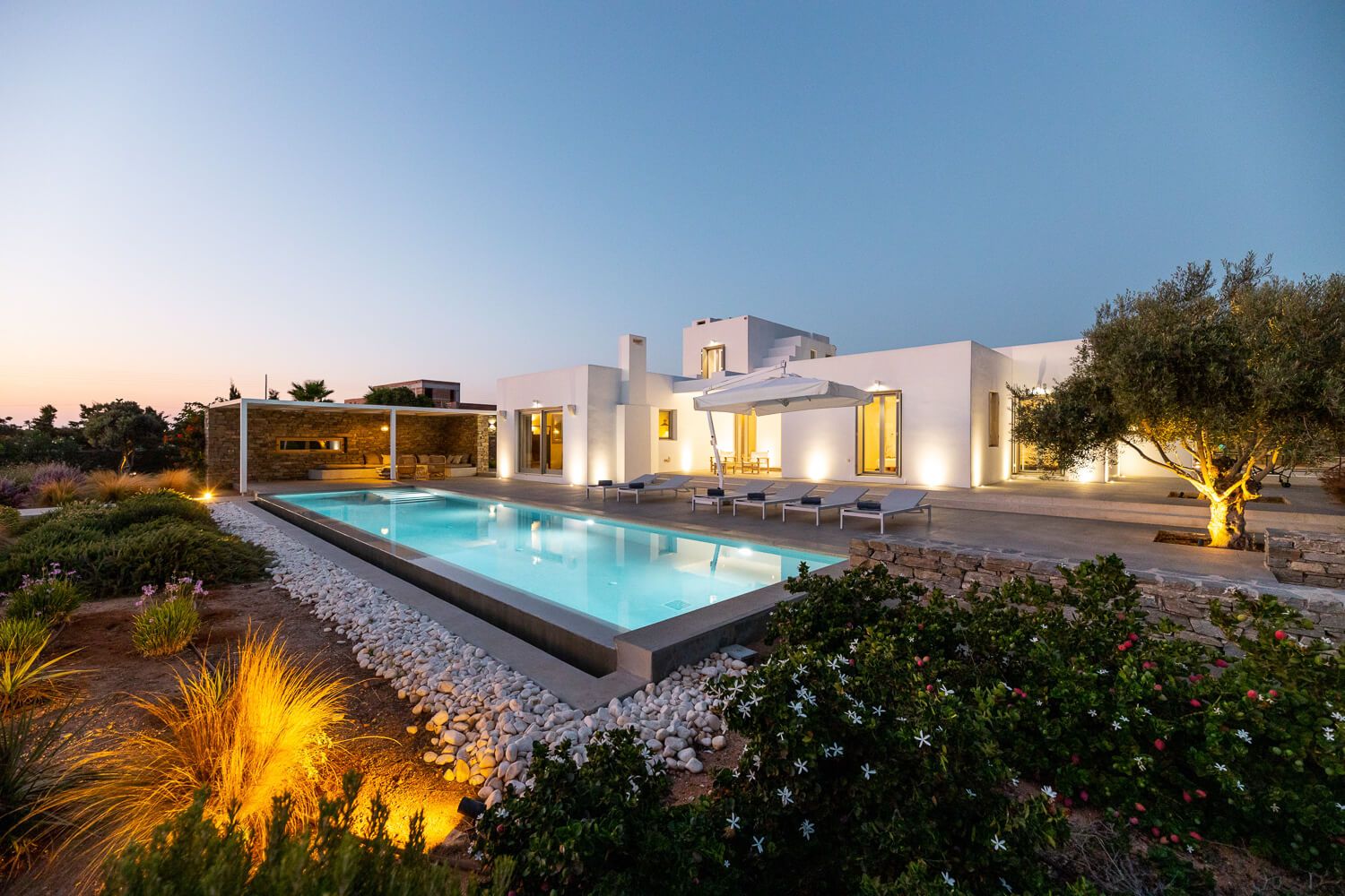 Modern villa with illuminated pool at dusk, surrounded by warm outdoor lighting in Paros.