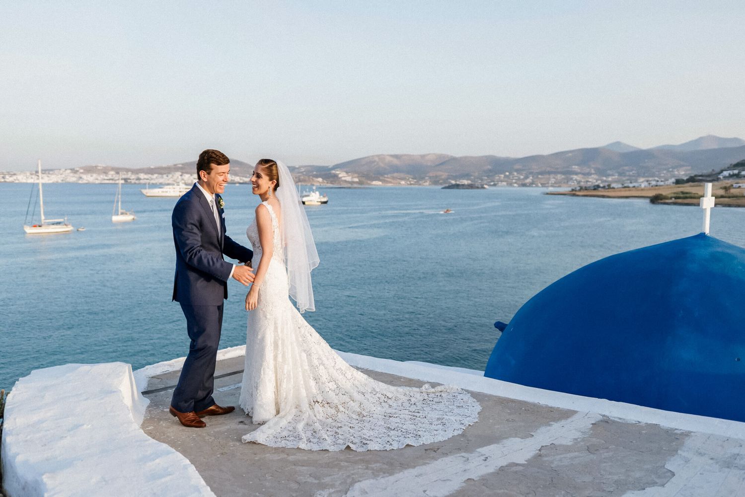 Bride and groom share a romantic moment on a white terrace overlooking the blue Aegean Sea in Paros, Greece.