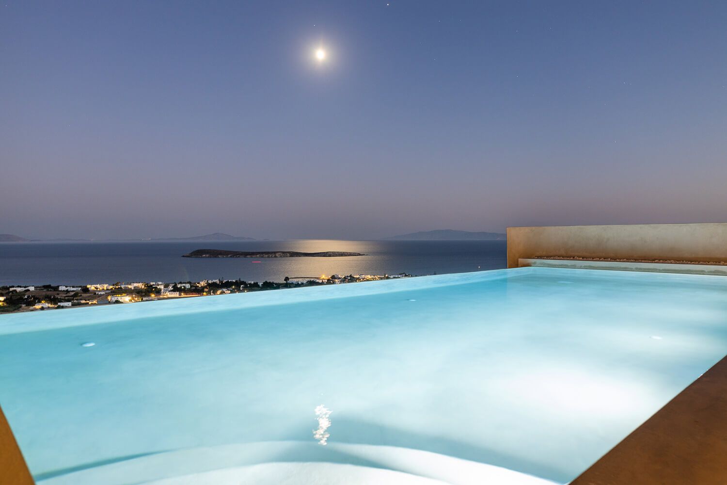 Illuminated pool overlooking the moonlit sea at dusk, with a serene island silhouette on the horizon