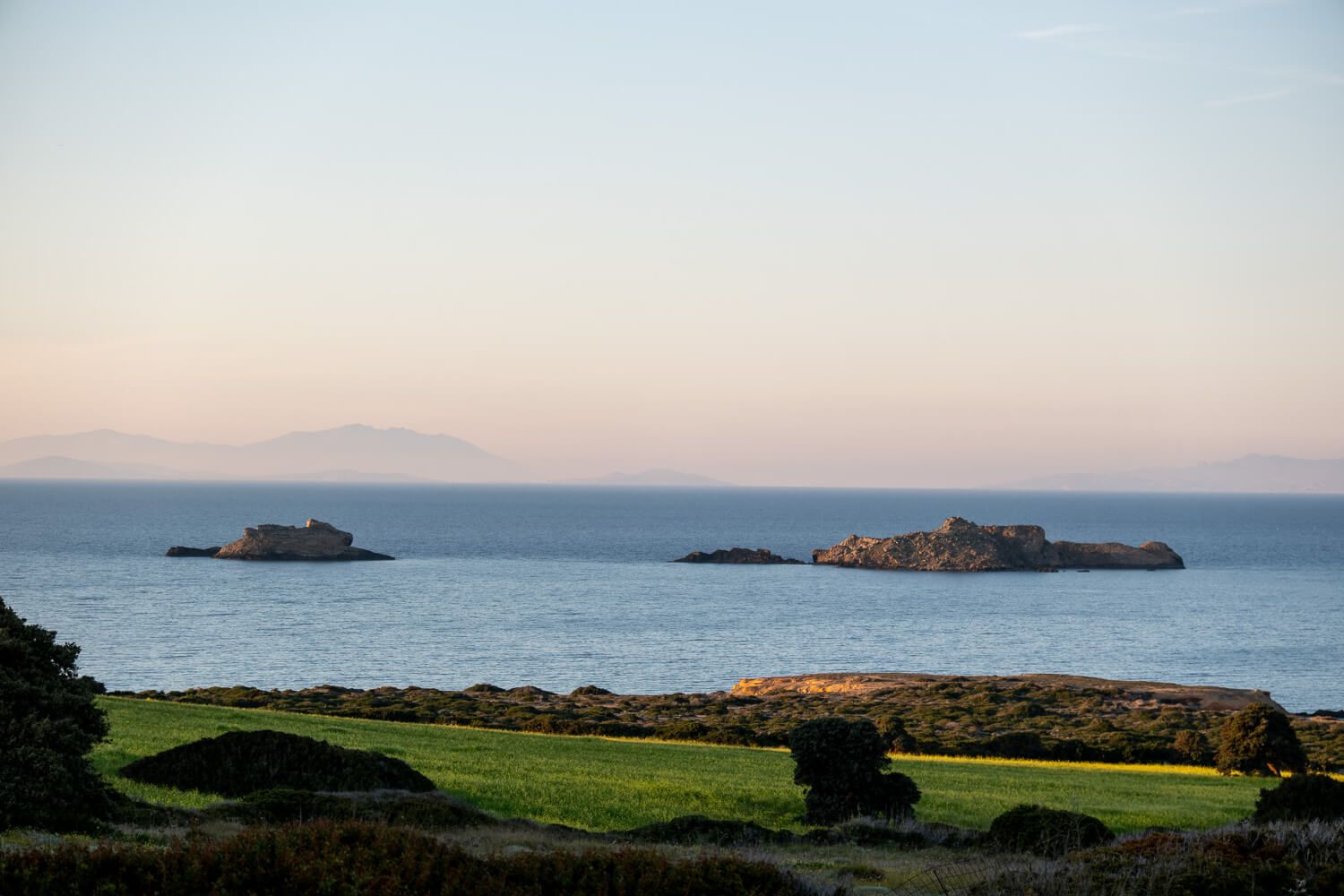 Panoramic coastal landscape showing rocky cliffs, green fields and distant islands along a misty ocean shoreline in Paros
