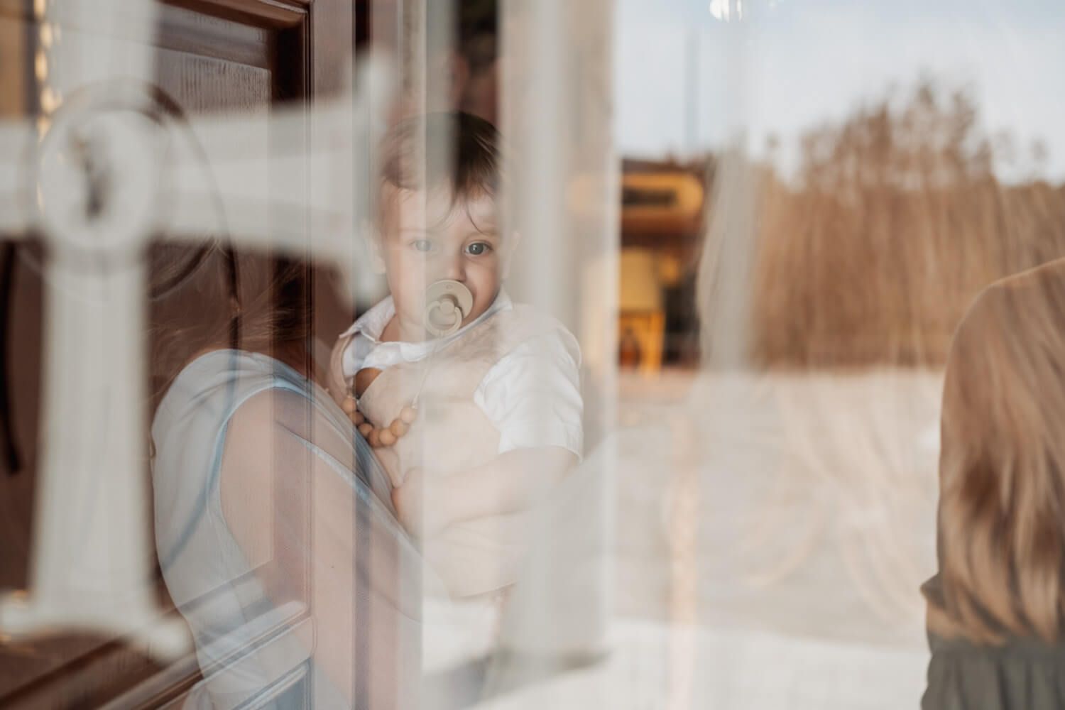 Baby in white christening outfit looking through a glass door.
