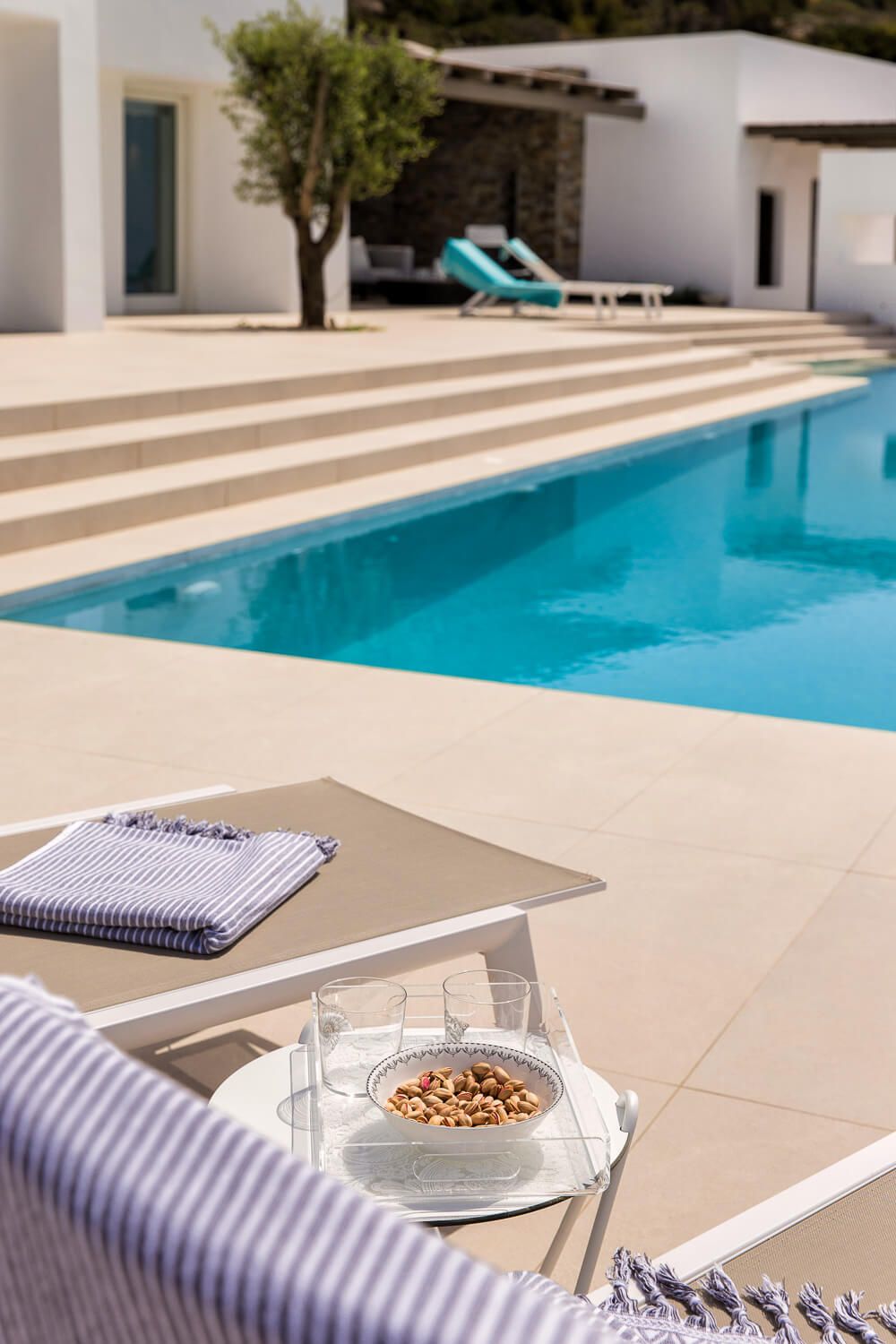 Luxury poolside lounge chair with snacks and drinks overlooking a modern swimming pool and white villa.