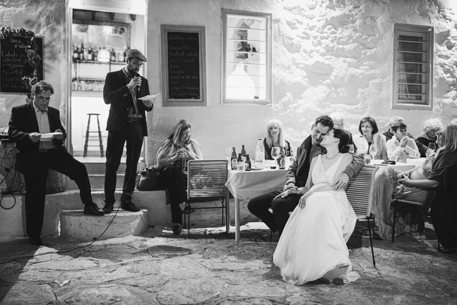 Black and white wedding reception scene with guests seated outdoors while someone gives a speech.