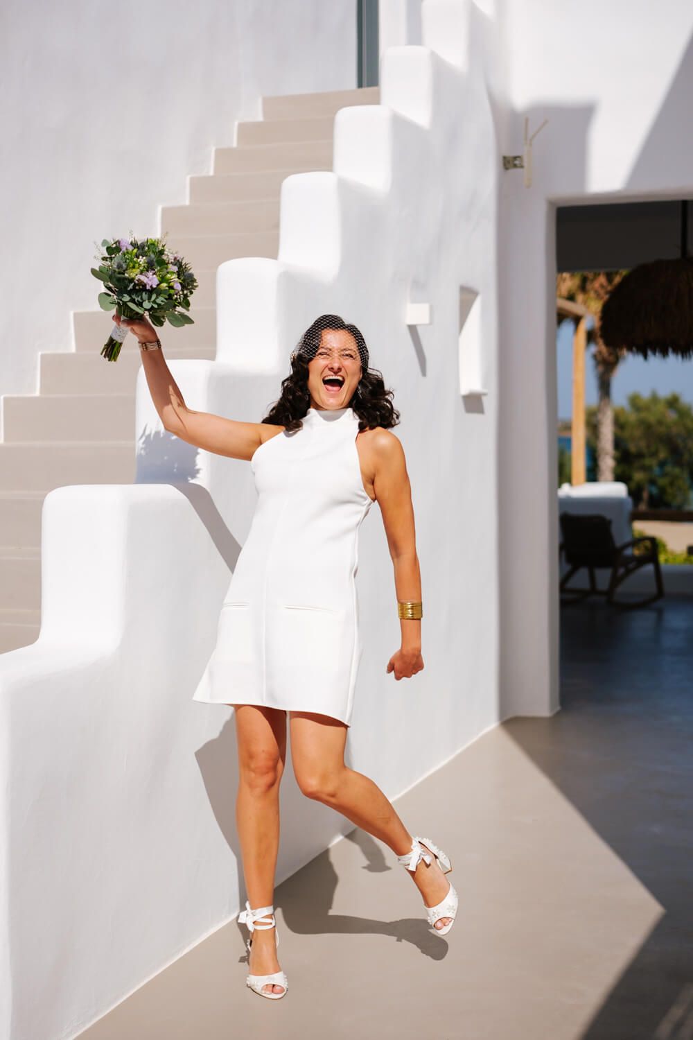 Joyful bride in short white dress holding bouquet aloft beside whitewashed staircase on her wedding.