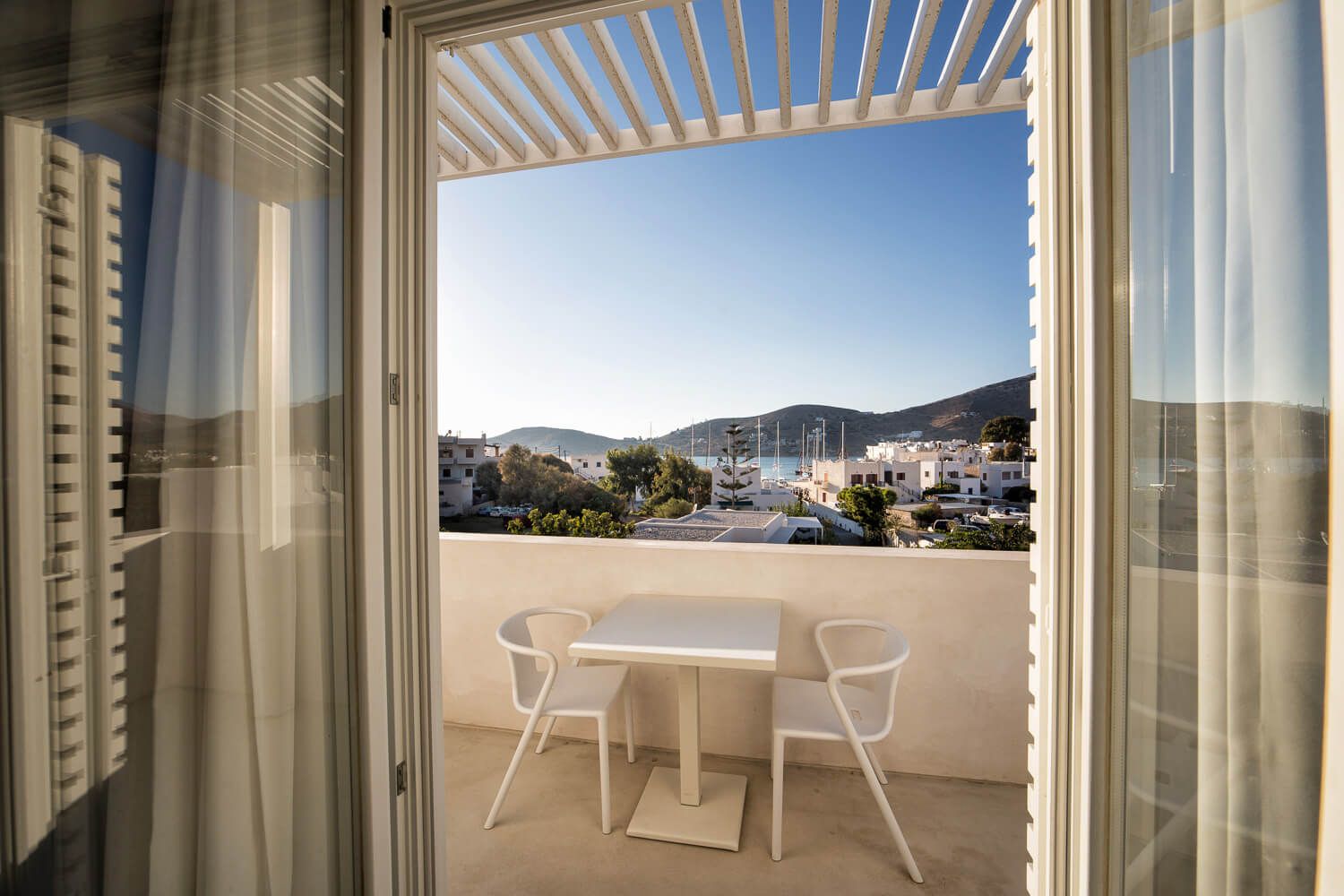 View through open doors to a sunny balcony with white furniture overlooking coastal mountains and buildings.